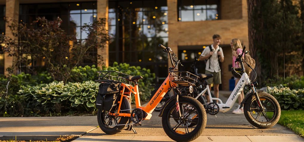 An orange Kingbull Voyager 2.0 cargo e-bike is parked in front of the academic building, with two students conversing behind it.