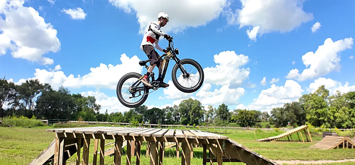 A cyclist on a fat tire e-bike mid-air, having just launched from a large wooden ramp, with a second ramp visible in the background against a bright blue sky with clouds.