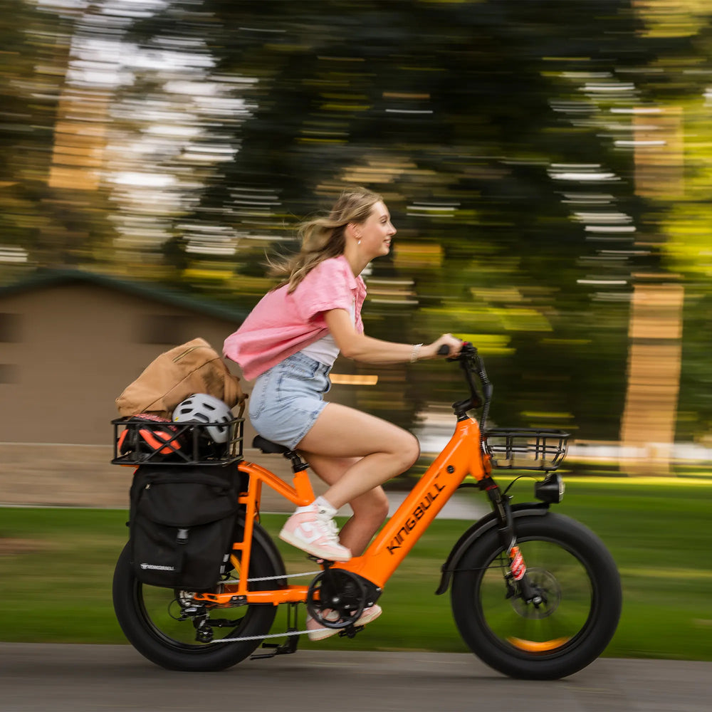Young woman riding orange Kingbull Voyager 2.0 electric bike with cargo racks and storage bags against blurred green trees background