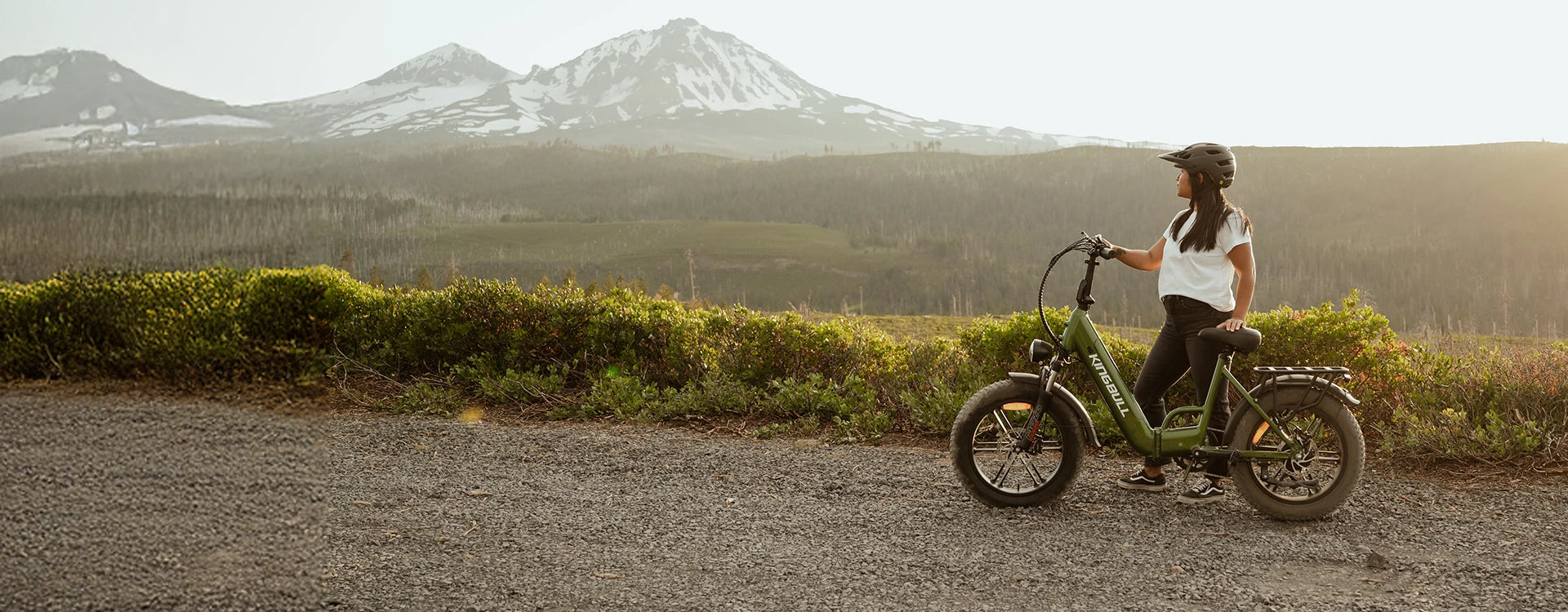 Woman wearing helmet riding green Kingbull Literider 2.0 electric bike on scenic mountain road with snow-capped peaks in background