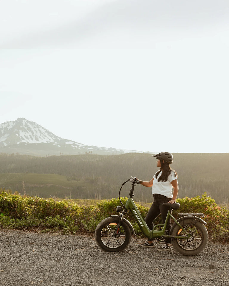 Woman wearing helmet riding green Kingbull Literider 2.0 electric bike on scenic mountain road with snow-capped peaks in background
