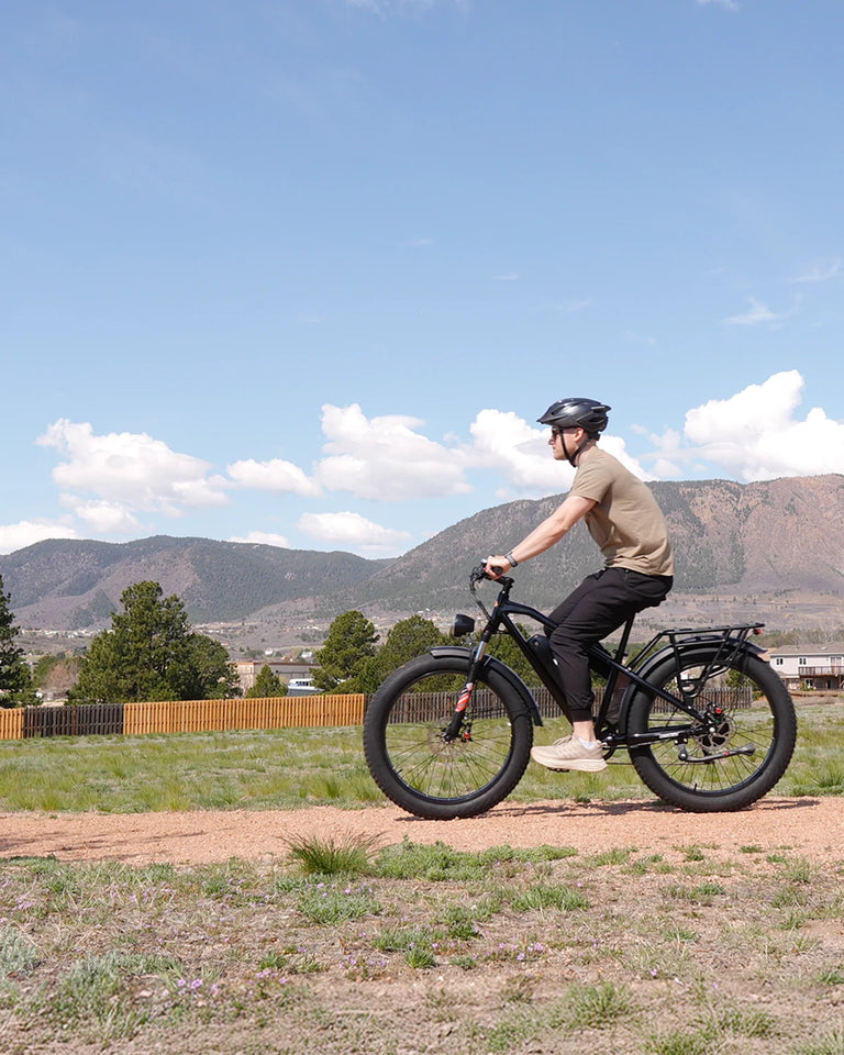 Man wearing helmet riding black Kingbull Hunter 2.0 electric fat tire bike on rural path with mountains in background