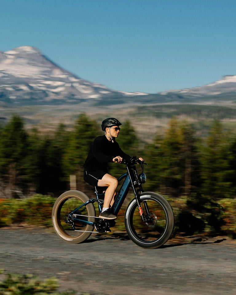 Man in black outfit riding blue Kingbull Rover 2.0 electric bike on paved road with snow-capped mountain and forest scenery