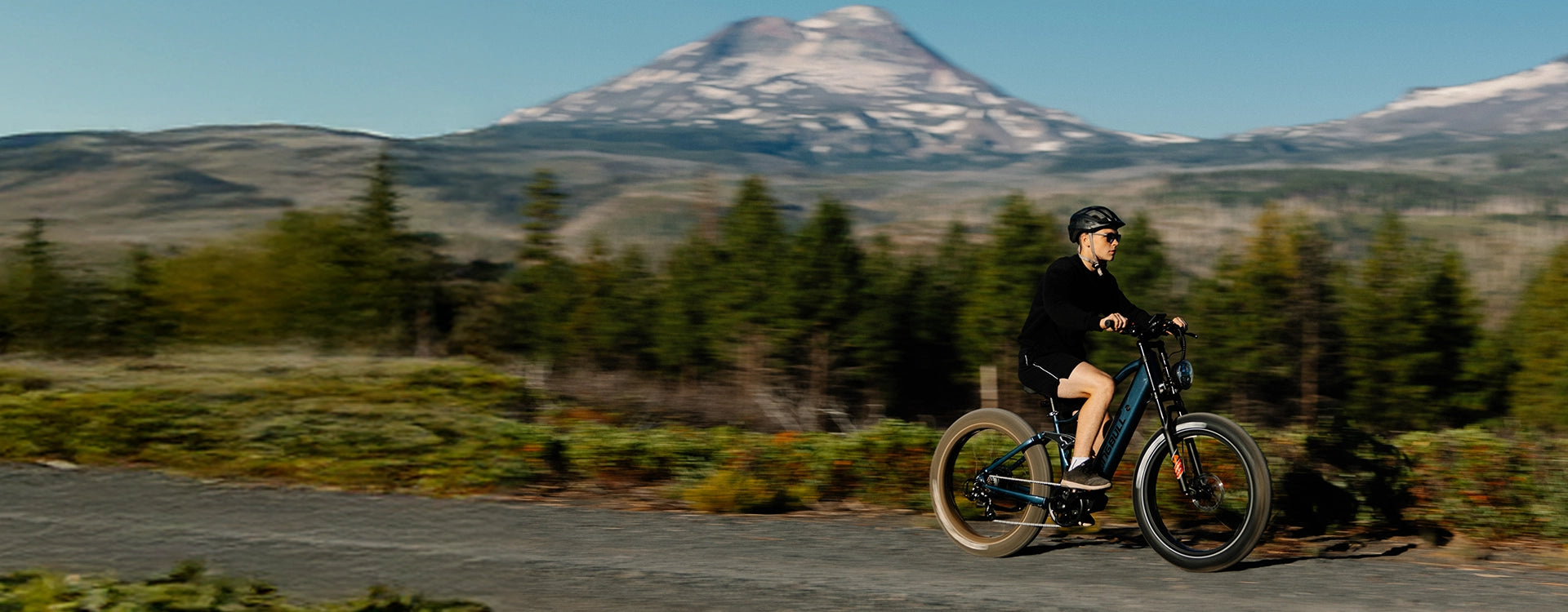 Man in black outfit riding blue Kingbull Rover 2.0 electric bike on paved road with snow-capped mountain and forest scenery